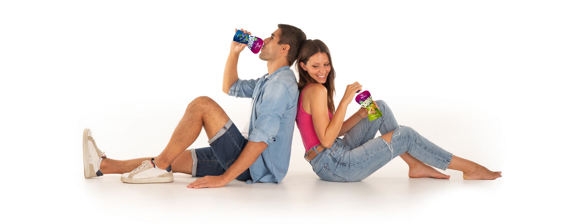 Ein junger Mann und eine junge Frau sitzen Rücken an Rücken auf dem Boden und trinken jeweils aus einer bunten Getränkepackung.