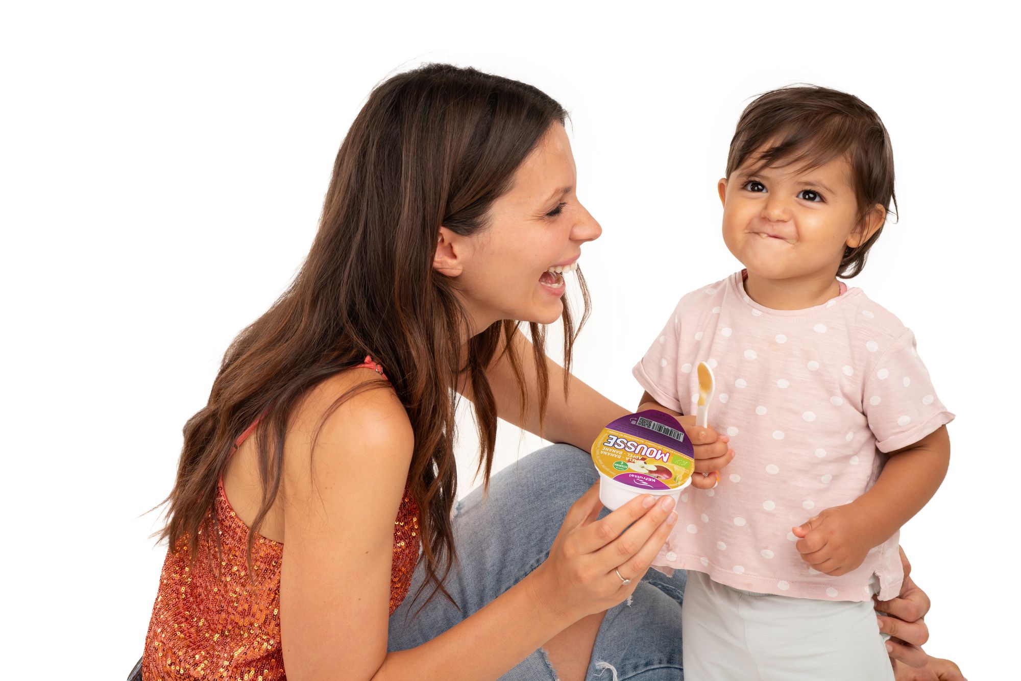 Eine Frau mit langem braunen Haar kniet neben einem kleinen Kleinkind, das ein Becher Mousse in der Hand hält.