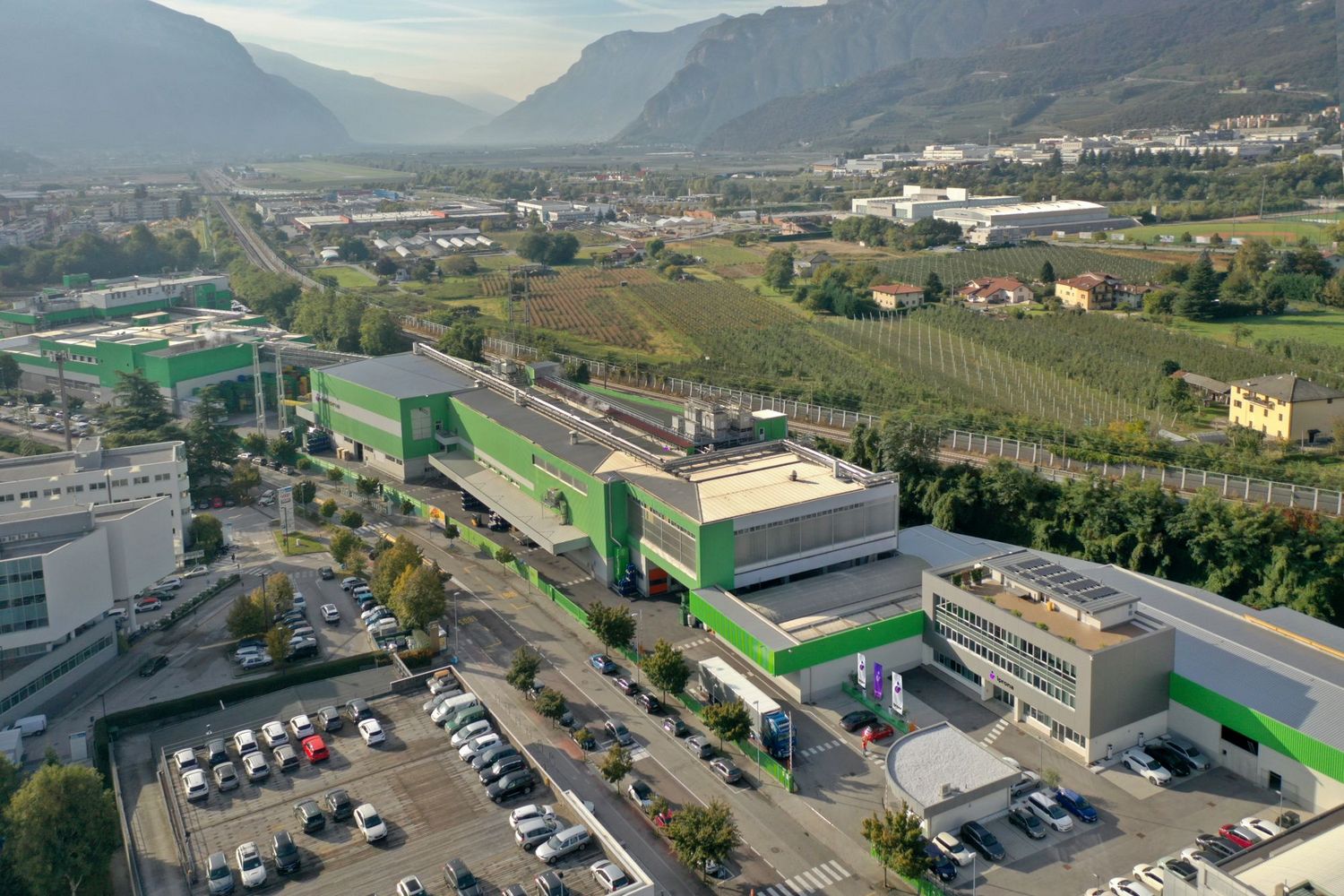 Luftaufnahme eines modernen Industrie- und Bürokomplexes mit markanten grünen Fassadenelementen, umgeben von einem Parkplatz und weiten landwirtschaftlichen Feldern vor einer Bergkulisse.
