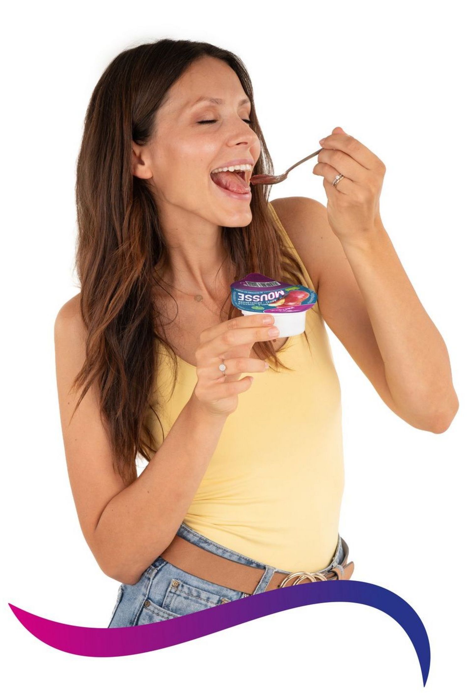 Eine junge Frau mit langen braunen Haaren und gelbem Top hält einen Becher Mousse in der Hand und probiert genussvoll einen Löffel davon.