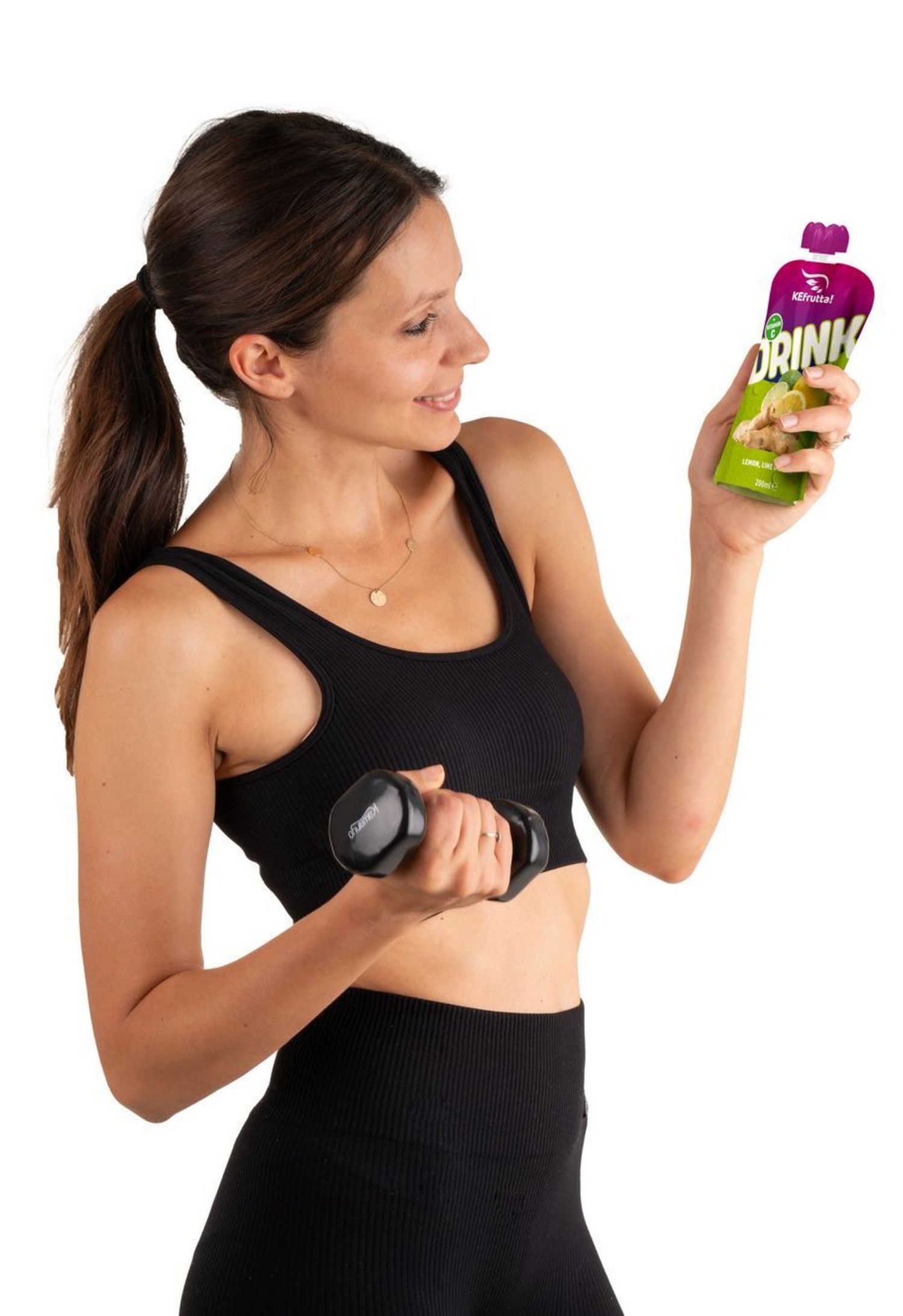 Eine junge Frau in Sportkleidung hält in einer Hand eine kleine Hantel und in der anderen ein Produktbeutel mit der Aufschrift Drink Zitronen-Limetten-Geschmack vor weißem Hintergrund.